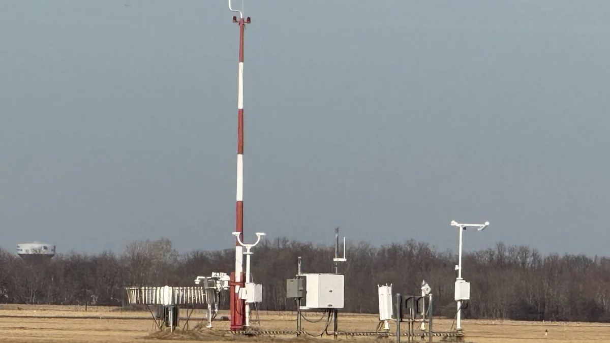 Automated weather station at an airport: a red and white wind sensor mast and white instrument boxes under a grey sky