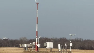 Estación meteorológica automática en un aeropuerto: mástil rojo y blanco con sensores de viento y cajas blancas de instrumentos bajo un cielo gris
