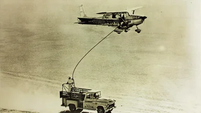 Foto antigua del Cessna 172 N9172B volando bajo sobre una carretera del desierto mientras sube una manguera de combustible desde una camioneta