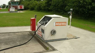 Self-service AVGAS 100LL fuel pump on a concrete apron at a small airfield, with a separate JET A-1 pump visible in the background near the control tower