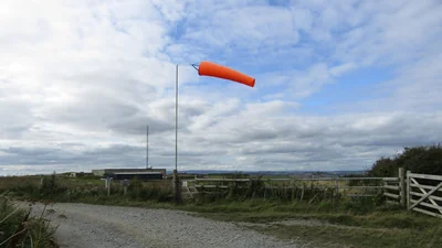 An orange windsock on a pole beside a gravel track and wooden gate at Bolt Head Airfield, under a cloudy sky
