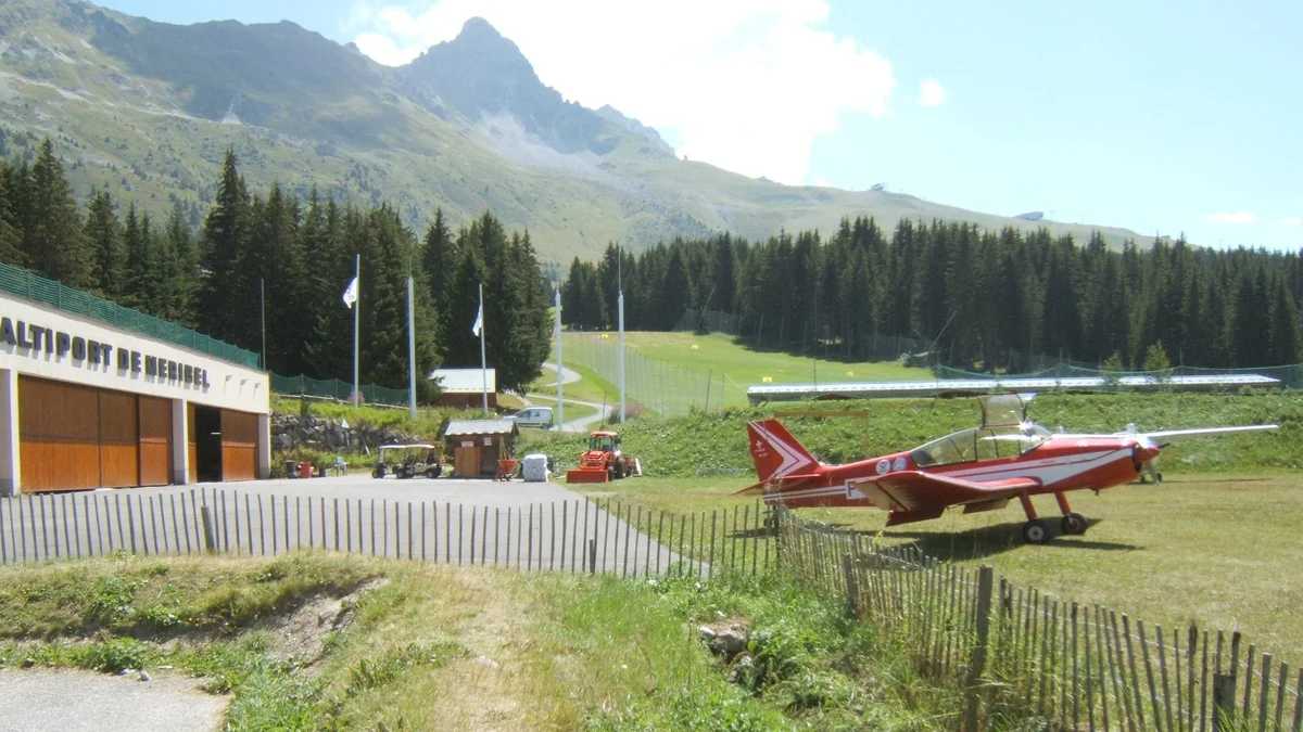 The Méribel altiport in the French Alps on a summer day, with a red light aircraft parked on the grass below steep mountain slopes
