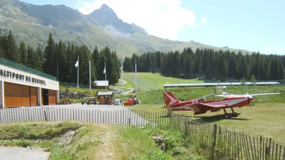 The Méribel altiport in the French Alps on a summer day, with a red light aircraft parked on the grass below steep mountain slopes