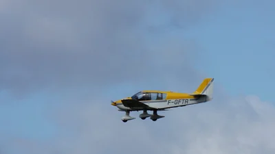 A yellow and white Robin DR400 light aircraft, registration F-GFTR, on final approach under a grey and blue sky