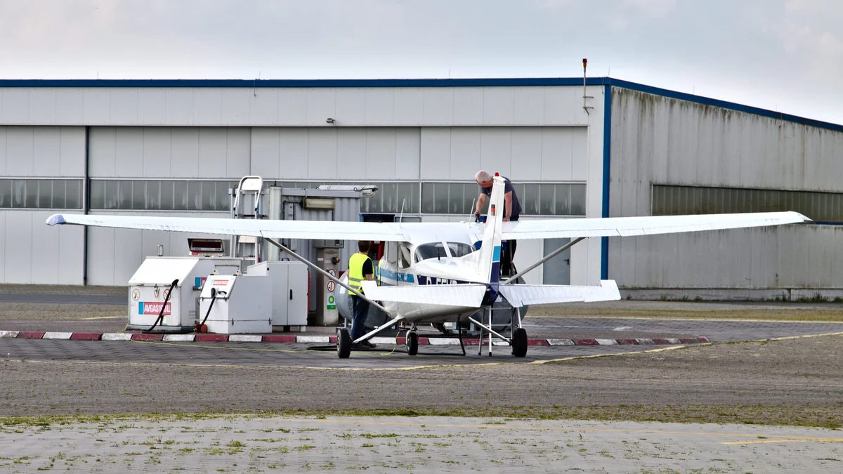 Cessna 172M being refuelled at a self-service fuel station on a grass airfield, with AvGas and MoGas tanks visible beside the wing