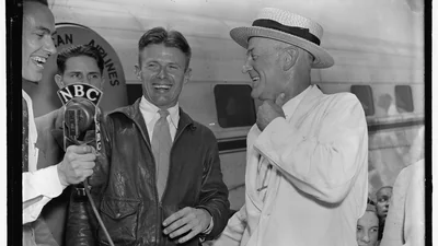 Foto en blanco y negro de 1938 de un sonriente Douglas Corrigan con chaqueta de cuero ante un micrófono de la NBC junto a un hombre con sombrero de paja, con un avión de línea detrás