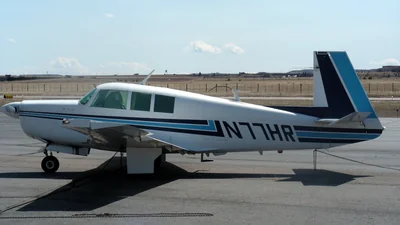 Un Mooney M20F blanco con franjas azules y negras estacionado en la plataforma de un aeropuerto bajo un cielo azul claro
