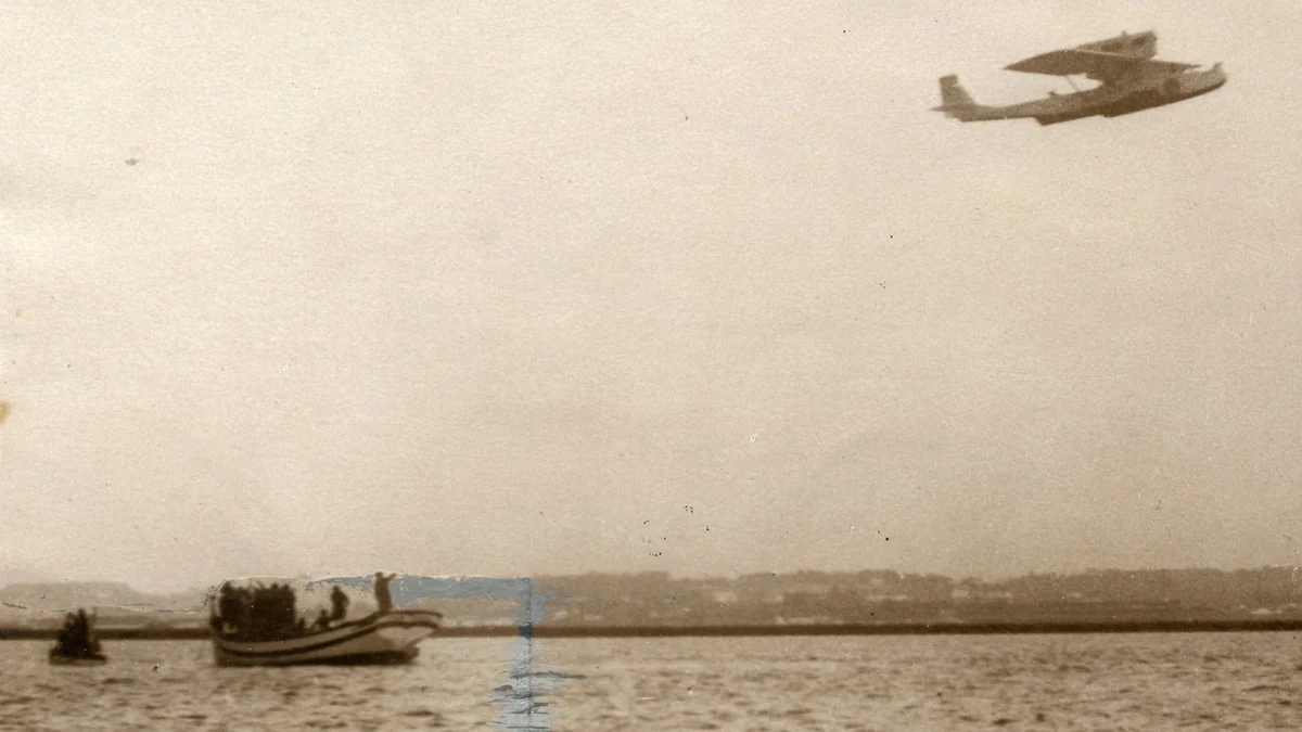 Sepia 1926 photo of the Dornier Wal flying boat Plus Ultra in flight low over the water, with small boats below