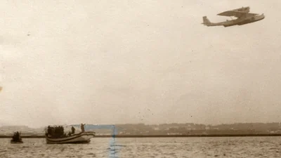 Foto en sepia de 1926 del hidroavión Dornier Wal Plus Ultra volando bajo sobre el agua, con pequeñas embarcaciones debajo