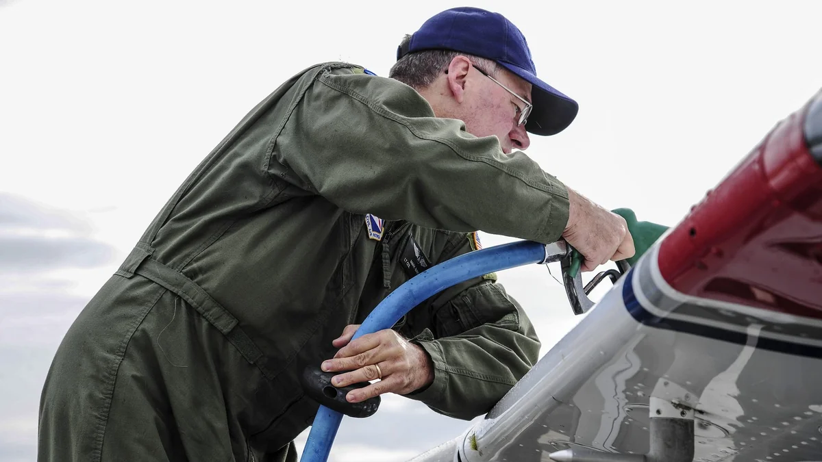 A pilot in a green flight suit and blue cap refuels the wing tank of a Cessna 172 with a blue fuel hose