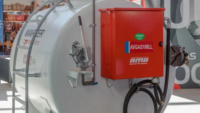 Depósito blanco de avgas expuesto en la feria AERO Friedrichshafen, con un armario surtidor rojo rotulado AVGAS100LL y una pegatina verde de Unleaded UL91